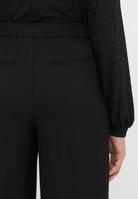 Pantalons noirs avec une texture lisse, dotés d'une taille élastique haute et de deux poches arrière. Le design est simple et ajusté.