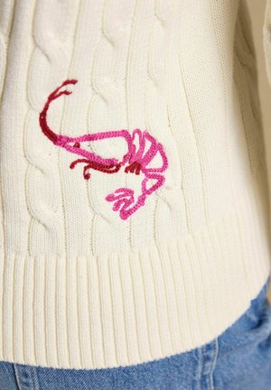 Kremo spalvos megztinis su pynimo raštu, ant kurio virš riboto apvado siuvinėtos rožinės ir raudonos krevetės, derinamas su mėlynais džinsais.
