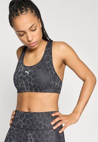 Μαύρο αθλητικό σουτιέν με γκρι λεοπάρ σχέδιο, κατασκευασμένο από ελαστικό υλικό. Διαθέτει σχεδιασμό racerback και μικρό λευκό λογότυπο μπροστά.
