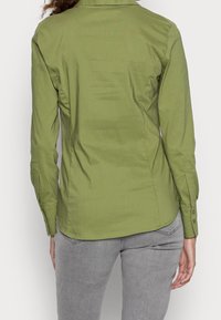 Camicia verde con bottoni, dal tessuto morbido, maniche lunghe e vestibilità su misura. Il retro presenta dettagli di cucitura e un orlo curvato.