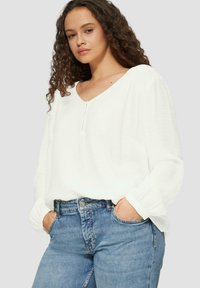 Witte, lange mouwen blouse met een V-hals en knoopdetail aan de voorkant, gecombineerd met hoog getailleerde blauwe jeans. Zachte, gestructureerde stof.