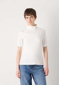 Hvit kortermet topp med høy krage, med en glatt tekstur og detaljer med glidelås ved halsen. Kombinert med blå denimjeans.