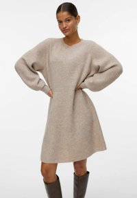 Frau mit zurückgebundenem Haar trägt ein beiges Strickkleid mit Puffärmeln und braunen Knielangen Stiefeln, steht mit den Händen in den Hüften.