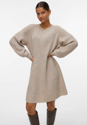 Femme avec les cheveux attachés porte une robe-sweater en tricot beige avec des manches bouffantes et des bottes marron jusqu'aux genoux, se tenant avec les mains sur les hanches.