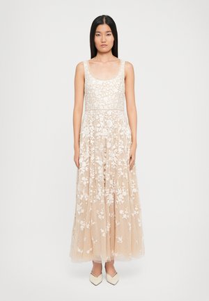 Nuori nainen seisoo yllään hihaton beige mekko, jossa on valkoinen kukkakuvioinen pitsikangas, ja teräväkärkiset valkoiset kengät yksinkertaista valkoista taustaa vasten.