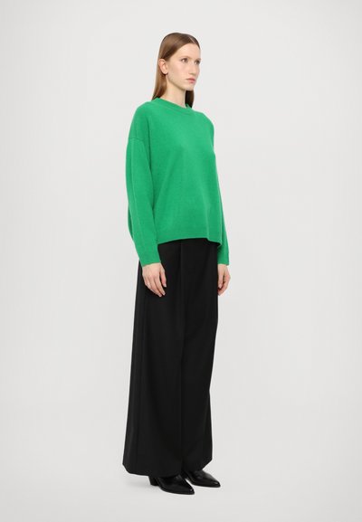Green knitted sweater with a relaxed fit, round neck. Paired with wide black trousers and black pointed shoes. Smooth texture, minimal design.