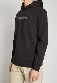 Personne portant un hoodie noir Calvin Klein et un pantalon beige, se tenant devant un fond gris clair uni.
