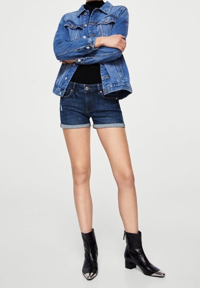 Personne debout, les bras croisés, portant une veste en jean bleue, un short en jean foncé, un haut noir et des bottines noires à bouts métalliques.