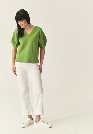 Nő zöld, rövid ujjú V-nyakú felsőt, fehér mandzsettás nadrágot és fehér tornacipőt visel, áll és enyhén mosolyogva lefelé néz semleges háttér előtt.