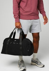 Man wearing gray shorts, maroon sweatshirt, sneakers, holding black duffel bag by its handles against gray background.