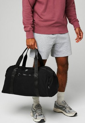 Mann in grauen Shorts, kastanienbraunem Sweatshirt und Sneakers, der eine schwarze Sporttasche an den Griffen vor einem grauen Hintergrund hält.