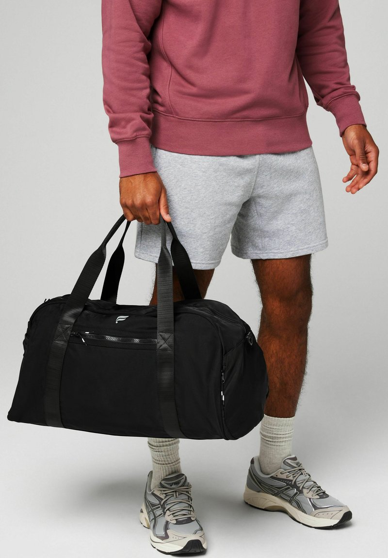 Man wearing gray shorts, maroon sweatshirt, sneakers, holding black duffel bag by its handles against gray background.