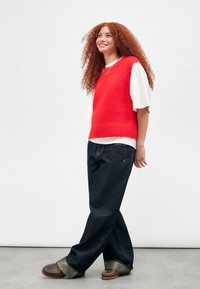 Junge Frau mit lockigem roten Haar, die einen roten ärmellosen Pullover über einem weißen Hemd, dunkle weite Jeans und braune Schuhe trägt, steht und lächelt.