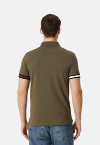 Polo de color verde oliva con cuello y mangas cortas, con acentos de rayas rojas y blancas en los puños. Textura suave similar al algodón.