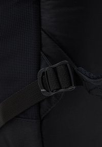 Černý batoh z látky s texturovaným mřížkovým vzorem, vybavený černou plastovou sponou a nastavitelnými popruhy pro bezpečné uzavření.