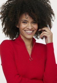 Roter gerippter Pullover mit hohem Kragen und einem silbernen Reißverschluss mit runder Ziehschlaufe. Der Stoff hat eine weiche, strukturierte Oberfläche.