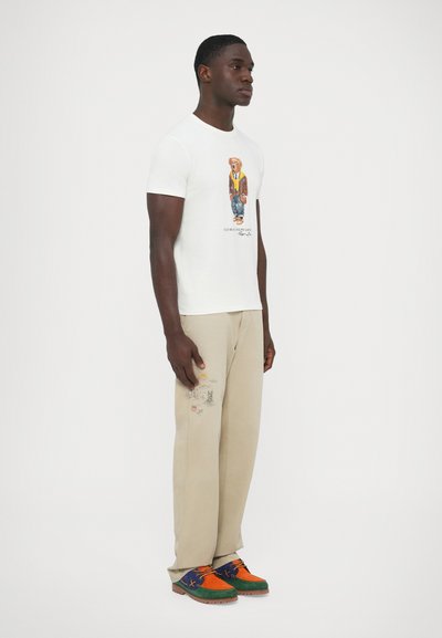 Camiseta branca com estampado gráfico de um urso com casaco, calças bege com detalhes bordados e sapatos multicoloridos com atacadores.