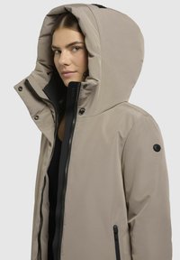 Beige gewatteerde jas met een waterafstotende stof, een zichtbare rits en zwarte accenten op de capuchon en zakken. Glad textuur, slim fit ontwerp.