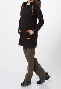 Abrigo largo negro con capucha ajustable, dos bolsillos delanteros y detalles de cuero; combinado con pantalones cargo oliva y zapatillas deportivas negras.