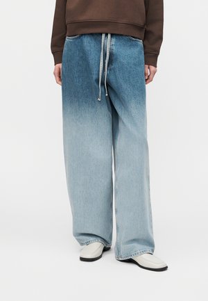 Personne portant un jean bleu délavé à jambes larges avec taille à cordon, un sweat-shirt marron et des chaussures blanches sur un fond blanc uni.