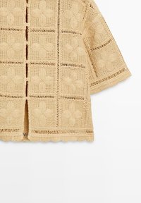 Beżowy krótki kardigan na szydełku z kwadratowymi wzorami kwiatowymi, falowanym obszyciem i małymi guzikami na środku przodu.