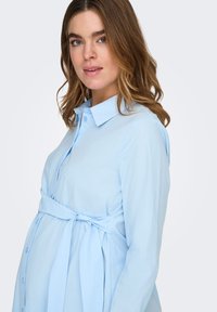 Γυναίκα με καστανά μαλλιά μέχρι τους ώμους, ντυμένη με ένα ανοιχτό μπλε πουκάμισο εγκυμοσύνης με κουμπιά, δεμένο πάνω από την κοιλιά της, κοιτώντας προς τα εμπρός με ελαφρύ χαμόγελο.