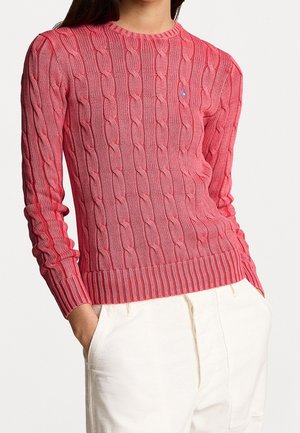 Vrouw draagt een roze kabelgebreide trui met lange mouwen en een witte broek, handen in de zakken, tegen een effen lichte achtergrond.