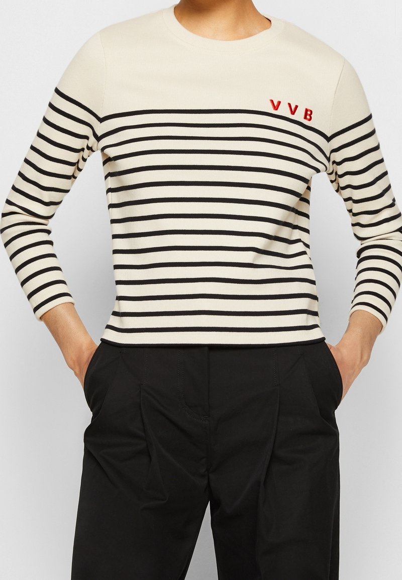Personne portant un haut blanc à manches longues avec des rayures horizontales noires et des lettres rouges "VVB", associé à un pantalon noir, mains dans les poches.