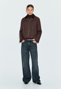 Braune cropped Jacke mit einem dunklen Kragen, goldenem Reißverschluss und zwei vorderen Taschen, kombiniert mit weit geschnittenen dunklen Jeans. Das Model steht aufrecht.