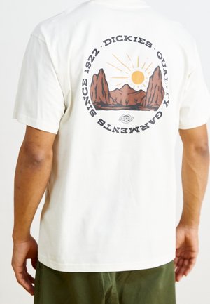 Osoba ubrana w białą koszulkę z grafiką pustynnego krajobrazu górskiego i słońca, z napisem „Dickies Quality Garments Since 1922” na plecach.