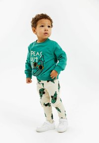 Mėlynos spalvos džemperis su „PEAS OUT“ grafika, spalvoti rankogaliai. Grietinėlės spalvos dryžuotos kelnės su žalių žirnių raštais. Baltos sportinės bateliai, stovėjimo poza, neutralus fonas.