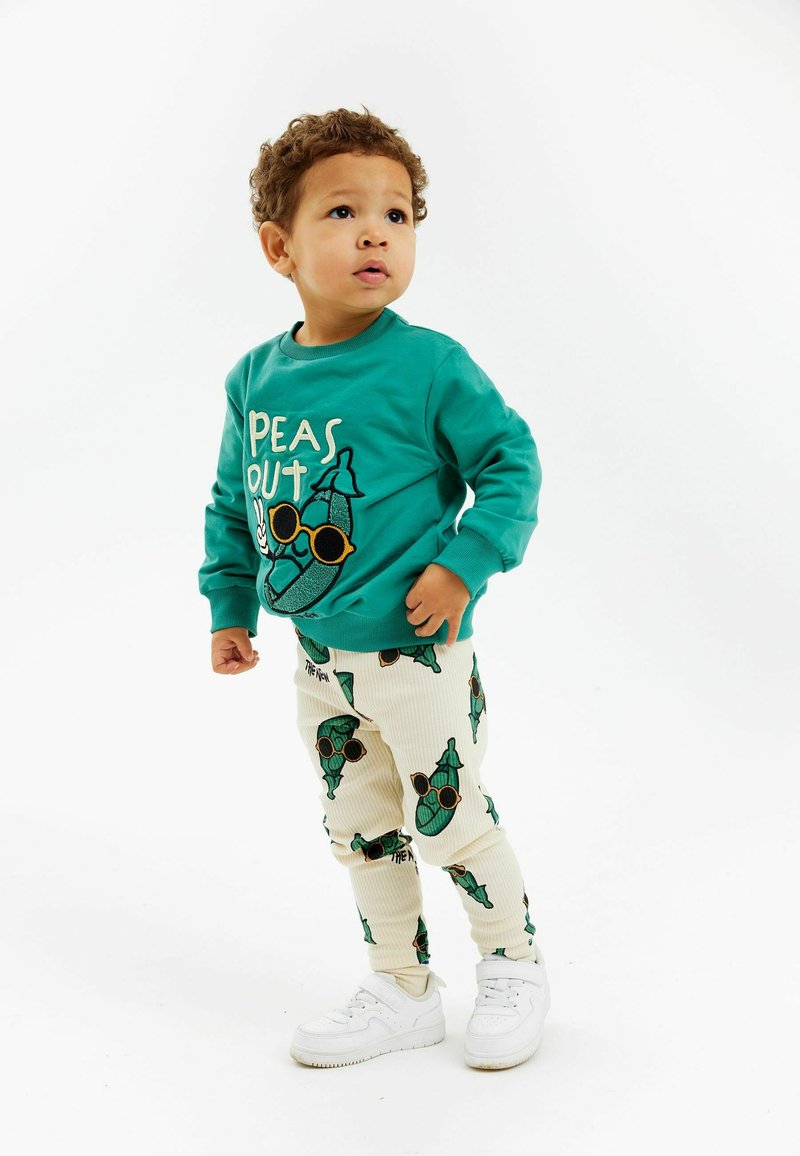 Mėlynos spalvos džemperis su „PEAS OUT“ grafika, spalvoti rankogaliai. Grietinėlės spalvos dryžuotos kelnės su žalių žirnių raštais. Baltos sportinės bateliai, stovėjimo poza, neutralus fonas.