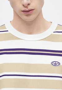 Μπλούζα με κοντά μανίκια και υφή βάφλας, που διαθέτει οριζόντιες ρίγες σε μπεζ, λευκό και μοβ. Περιλαμβάνει ένα μικρό λογότυπο στο αριστερό στήθος.