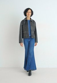 Grå lærjakke med krage, kombinert med en denimtopp og vide blå jeans, stående foran en nøytral bakgrunn. Svart ankelstøvler.