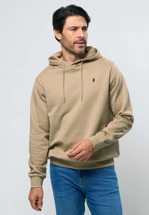 Vyras dėvi gelsvą gobtuvą megztą džemperį ir mėlynas džinsines kelnes, žvelgia į dešinę su neutraliu veido išraiška, fonas – paprastas.