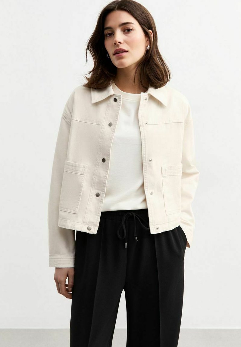 Veste en denim crème avec un col, fermeture à boutons sur le devant et deux poches poitrine, portée sur une chemise blanche et un pantalon à cordon noir.