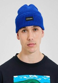 Berretto di lana blu lavorato a maglia con un motivo testurizzato e un'etichetta rettangolare nera. Indossato da una persona con i capelli corti e una maglietta scura sotto.