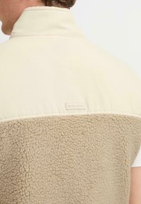 Beige Fleece-Weste mit einem cremefarbenen oberen Abschnitt. Verfügt über einen Stehkragen und einen strukturierten unteren Teil. Das Markenetikett ist auf der Rückseite sichtbar.