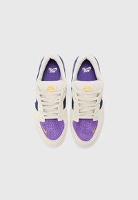 Les baskets Nike SB présentent un dessus crème, une capuche de orteil violette, des accents de côté bleus marine et un logo jaune sur la languette et le swoosh.
