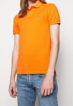 Orange Polo-Shirt aus Baumwolle mit einem Kragen, kurzen Ärmeln und einem blauen Logo auf der Brust. Getragen mit hellen Jeans.