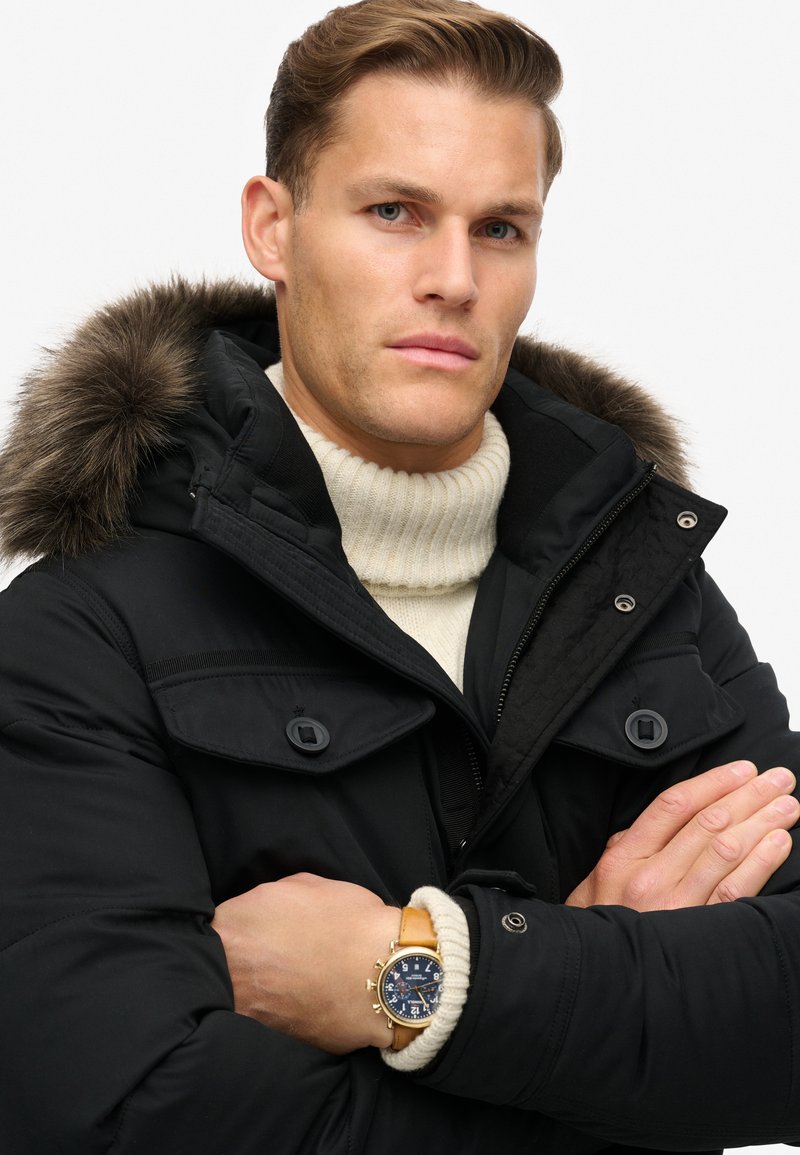 Zwarte gewatteerde jas met een gevoerd capuchon van bont, gelaagd over een crèmekleurige ribbeltrui. Bevat knopen en een horloge met een leren band.