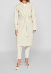 Abrigo color crema con cinturón, cuello chal, mangas largas y bolsillos delanteros, combinado con pantalones claros y zapatos de tacón con punta afilada.