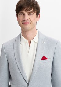 Hellgrauer Blazer mit strukturiertem Finish, ausgestattet mit einem roten Einstecktuch und einer silbernen Reversnadel. Darunter ein cremefarbener, gestrickter Poloshirt.