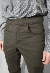 Pantaloni verdi in cotone dal taglio aderente, con tasche frontali e chiusura con bottone, abbinati a una camicia blu e bianca con motivo a fantasia.