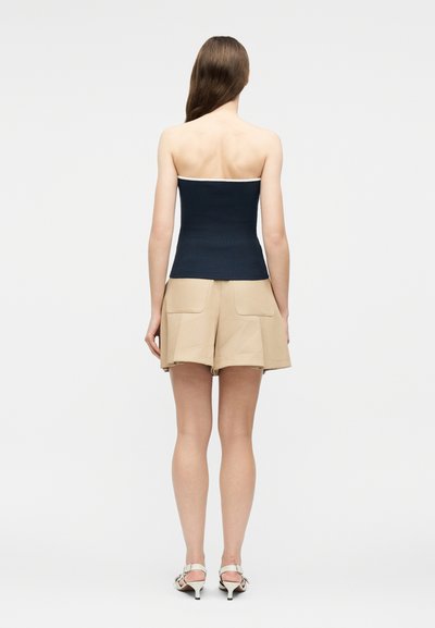 Γυναίκα με μακριά καστανά μαλλιά που φοράει μπλε σκούρο τοπ χωρίς τιράντες, μπεζ σορτς με ψηλή μέση και μεγάλες πίσω τσέπες, και λευκά πέδιλα με τακούνι, όρθια σε άποψη από πίσω.