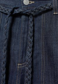 Tissu en jean bleu foncé avec des coutures beiges visibles, un bouton argenté et une ceinture tressée en jean passée dans les passants de ceinture.