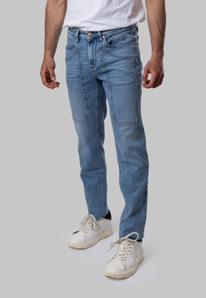 Uomo che indossa jeans azzurri chiari con tasche applicate davanti e sneakers bianche, in piedi davanti a uno sfondo grigio.
