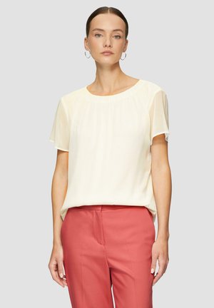 Kurzärmlige, crèmefarbene Bluse aus leichtem Stoff mit gerafftem Ausschnitt, kombiniert mit korallenfarbenen Hosen im entspannten Schnitt.