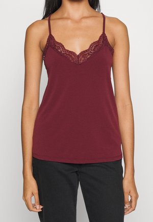 Vrouw draagt een mouwloos bordeauxkleurig hemdje met kanten rand en zwarte broek, staand tegen een effen lichte achtergrond.