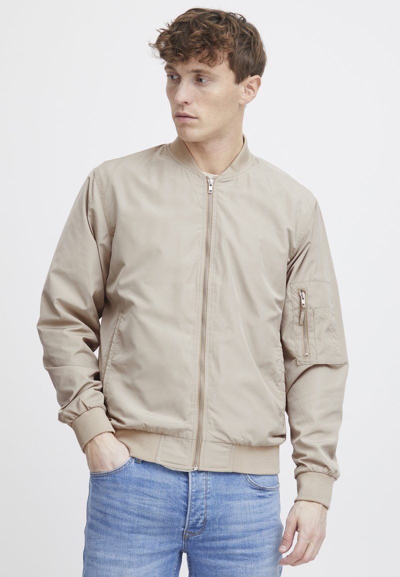Beige bomberjakke laget av glatt stoff, med frontglidelås og ribbede mansjetter. Inkluderer en glidelåslomme på venstre arm.